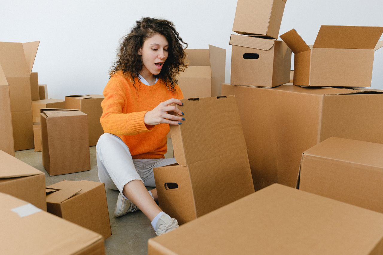 Amazed young female with curly hair in casual clothes sitting on floor and unpacking carton boxes with opened mouth after relocation